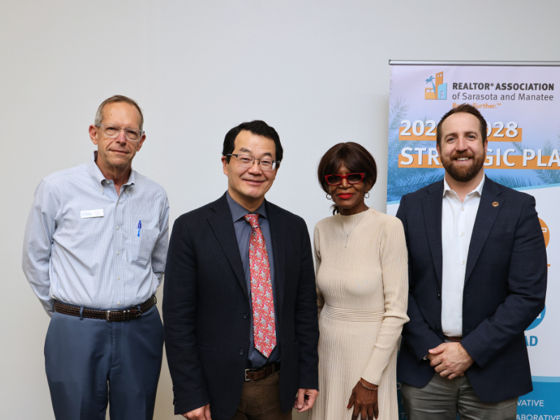 Four professionals stand together at the REALTOR® Association of Sarasota and Manatee in front of a RASM strategic plan banner.